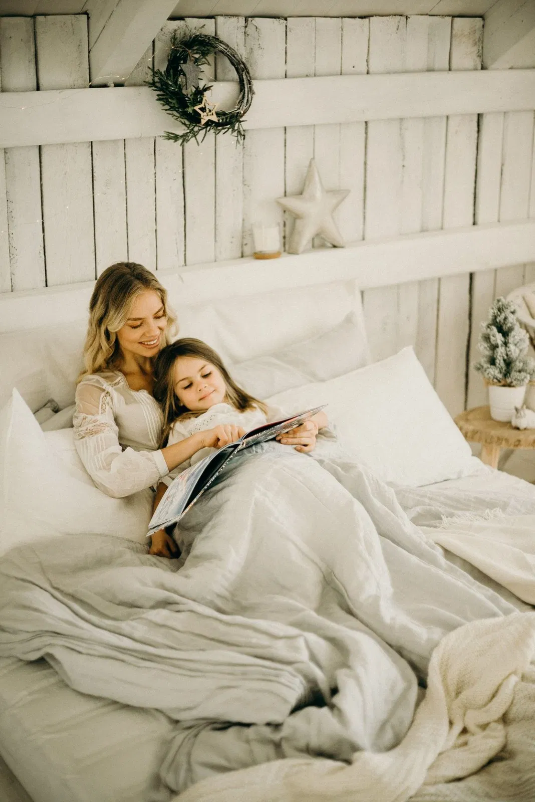 Mãe e filha loiras a ler juntas numa cama confortável.